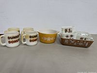 Full lot showing four Dunkin' Donuts mugs, yellow Pyrex bowl, and four small nesting Pyrex dishes arranged in a row.