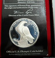 Silver proof coin featuring a bald eagle with 'United States of America One Dollar' text, certified by Donna Pope, Director of the United States Mint, with Olympic coin holder text visible.
