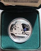 Close-up of silver coin featuring ship Santiago with '1774-1999' and 'CANADA DOLLAR' inscriptions in protective case inside green box.