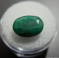 Close-up of the 5.8CT green emerald gemstone showing its cut, color and surface details on a white cushion inside a clear round container.