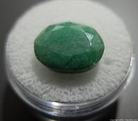 Close-up of the green emerald gemstone showing the faceted oval shape on white cushion inside clear round container.