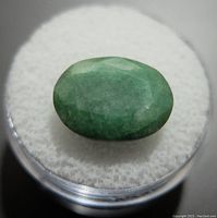 Close-up displaying the oval green emerald gemstone placed on white cushioned surface inside a clear round container.