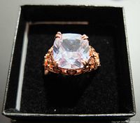 Front view of the 925 stamped silver fashion ring with a large cushion-cut clear gemstone in a rose gold-tone filigree setting inside a black box.