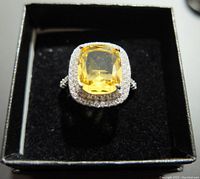 Top-down photo showing silver ring with large rectangular yellow gemstone center and two rows of smaller clear stones around it. Ring set in black display box.