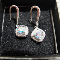 Pair of gemstone style earrings in black jewelry box, showing square iridescent stones surrounded by clear rhinestones, set in silver-tone metal with fish hook backs.
