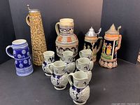 Full lot showing 11 German steins, jugs, and mugs, various sizes from 5 to 13.5 inches tall, including musical base stein and blue circle relief stein