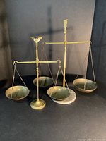 Pair of brass balance scales on dark background; larger with horn player finial, smaller with eagle finial on stone base