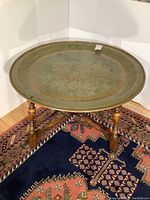 Copper top coffee table showing top etched decorative pattern, round copper surface with patina and wooden legs with cross support base on traditional patterned rug.