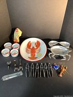 Photo of full lobster and crab entertaining set on neutral backdrop showing platter, butter cups, pewter crab dish, cutlery, skewers, ornaments, dish towel and cocktail glass.