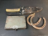 Knife with stag handle laid on top of the leather sheath, two horseshoes and metal box visible as lot contents
