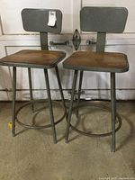 Front view of two vintage grey metal drafting stools with wooden seats and adjustable backs.