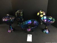 Group photo of six dark blue carnival glass pieces with iridescent finish showing multiple shapes and pedestal bases, grape cluster patterns.