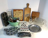 Wide shot showing entire lot of vintage kitchen items including glass butter dishes, metal plate, wooden cutting boards, utensils, grater, muffin pan, and recipe book.