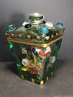 Front view of green square art glass jar with decorative glass balls on lid and gold painted edge accents, signed VA
