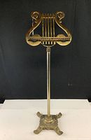Full view of the brass music stand, showing the lyre-shaped top, straight central column, and ornate base with three detailed ornamental legs.