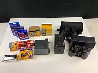 Photo showing all items in the lot arranged on table including cameras, binoculars, film packs, and camera box and flash.