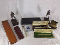 Full lot display showing all items: two Cross pens in cases, Proebster Technica case, Augusta brass lift lighter, two metal figurine desk sculptures, two leather pen cases, glass pen holder, small silver-toned box