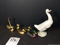 Group photo of all seven figurines: one white porcelain duck (Nao by Lladro), one painted cast iron mallard, two pairs of brass ducks and ducklings, and one brass swan, on black background.
