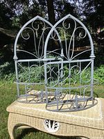 Two gray-painted iron planters with arched backs and scroll designs displayed outdoors on wicker table.