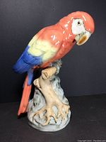 Front angle view of the ceramic parrot perched on a branch showing the coral red head, yellow shoulders, and blue wings and tail feathers.
