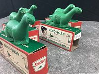 Five green dinosaur-shaped soaps embossed with 'DINO' and the original Sinclair Dino Soap Castile boxes behind them on a table.