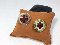 Two vintage lapel pins displayed on a brown cushion. Left pin is circular gold-tone frame with red enamel cross and engraved text. Right pin is round with red cross and yellow ring with text.