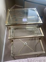 Three gold metal and glass nesting tables, stacked together showing varying sizes and clear glass tops with some corner chips.