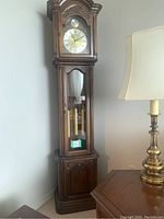 Full view of tall wooden grandfather clock with glass door showing clock face and pendulum weights inside. Clock located beside wooden side table and lamp.