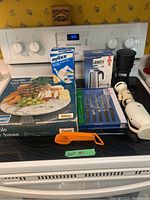 Image of kitchen items on stove including Bialetti Musa espresso maker box, Zyliss cheese slicer box, black drip coffee maker, Unger electric hand blender, steak knife set, marble Lazy Susan box, orange measuring spoons and salt shaker.