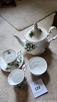 Photo showing teapot with lid, teacup, saucer, creamer, sugar bowl lid, and small bowl arranged on a carpeted floor.