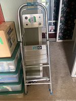 Front view of aluminum step ladder folded and leaned against a wall near plastic storage bins and cardboard box.