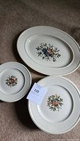 Three pieces including one large oval serving platter and two round plates with floral designs and ribbed borders, laid on carpet.