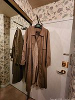 Front view of beige women's trench coat hanging on hanger on door, showing collar, buttons, and interior checkered lining.