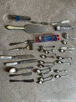 Photo showing mixed silver flatware including spoons, forks, knives, serving utensils, and boxed spoon on carpet background.