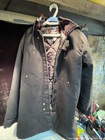 Full view of the dark canvas men's winter coat with hood hanging on a hanger, showing front zipper and pockets.