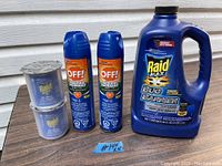 Photo showing a large blue plastic bottle of Raid Max Bug Barrier, two tall aerosol cans of OFF! Deep Woods insect repellent, and two smaller blue Raid Dry Fogger insecticide cans arranged on wooden surface.