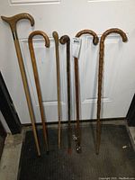 Six antique wooden canes standing against a door, showing full length and different handle shapes and wood textures.
