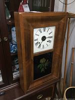 Front view showing rectangular wooden ogee frame with white clock face and Roman numerals above floral decorated black glass panel below.