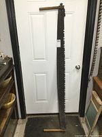 Full length view of the antique saw showing the entire blade and wooden handles at either end, positioned against a door for scale.