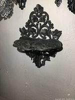 Front and side views of a cast iron wall shelf with intricate leaf and flower designs mounted on a wall.