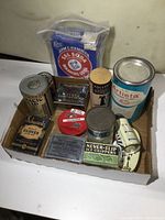 Photo of various vintage household tins and boxes in a cardboard tray, including Arm & Hammer Sal Soda Washing Soap in plastic bag, Artista cleaner, Schwartz Cloves, Paterson's Toothpicks, Never-Slip Ice Grippers, and other assorted tin cans.