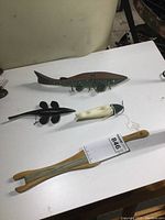 Three handmade weighted wood fish decoys of different sizes and a wooden jigging stick on a white surface.