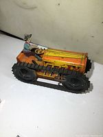 Side view of vintage windup metal tractor toy showing lithographed yellow/orange body, black rubber tracks and seated driver figure in green and red attire.