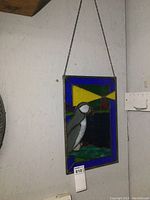 Front view of stained glass panel showing puffin and lighthouse design, hung by metal chain against wall.