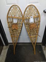 Full front view of both wooden snowshoes displayed side by side on a door background, showing overall shape and construction