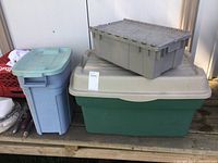 Three plastic totes including an extra large green tote with beige lid, a medium blue tote with lid, and a medium gray tote with hinged lid on top of the green tote.