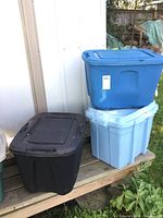 Three large plastic storage totes; black tote on the left, blue tote stacked on light blue tote on the right, located outdoors.