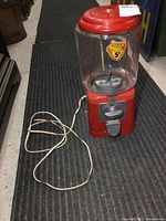 Full view of the vintage Acorn candy vending machine showing the glass container, red metal base and top, and attached power cord on a carpeted floor.