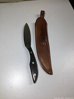 Full view of cutter with wooden handle next to brown leather sheath on white surface.