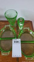 Two green depression glass divided dishes with floral etching and handles, accompanied by three green etched glass drinking vessels on a wooden surface.